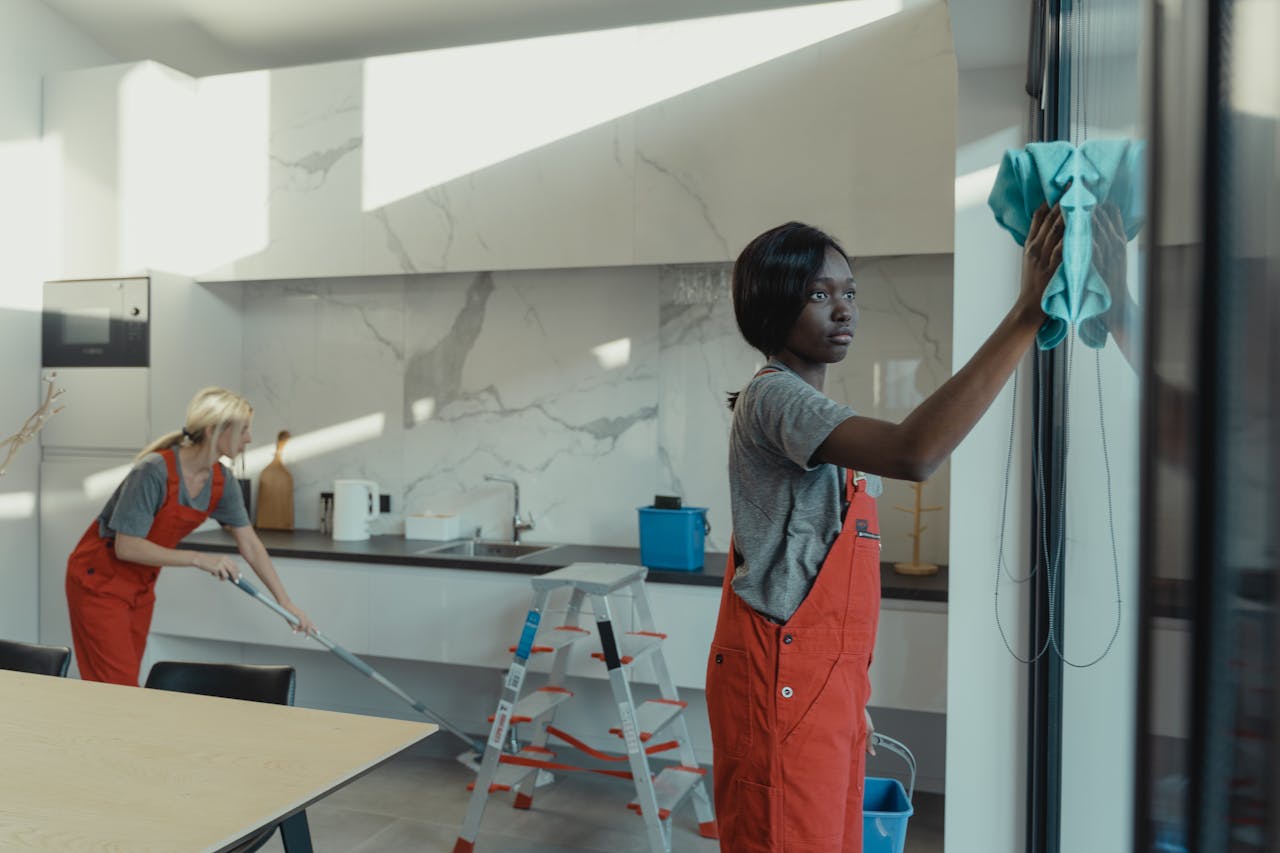 expertise-01 Two women cleaning a modern kitchen, wiping glass surfaces and mopping the floor.